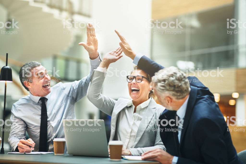 Ecstatic group of mature work colleagues high fiving together while having a meeting at a table in the lobby of a modern office building